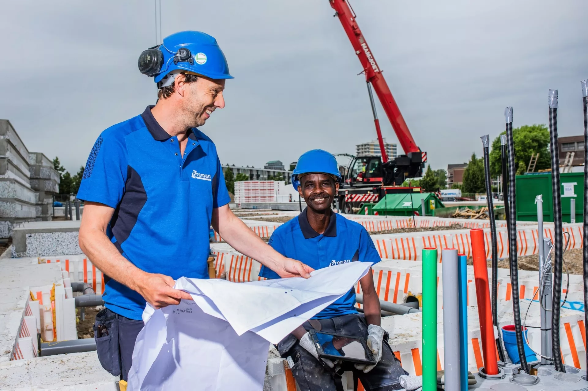 Twee Breman-monteurs op een bouwplaats bekijken samen een bouwtekening. Op de achtergrond staat een kraan en zijn installatiematerialen zichtbaar.