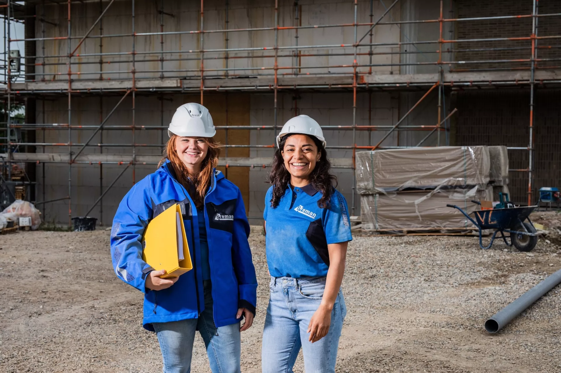 Twee Breman-medewerkers met veiligheidshelm op een bouwplaats, lachend en in werkkleding, voor een steiger en bouwmaterialen.