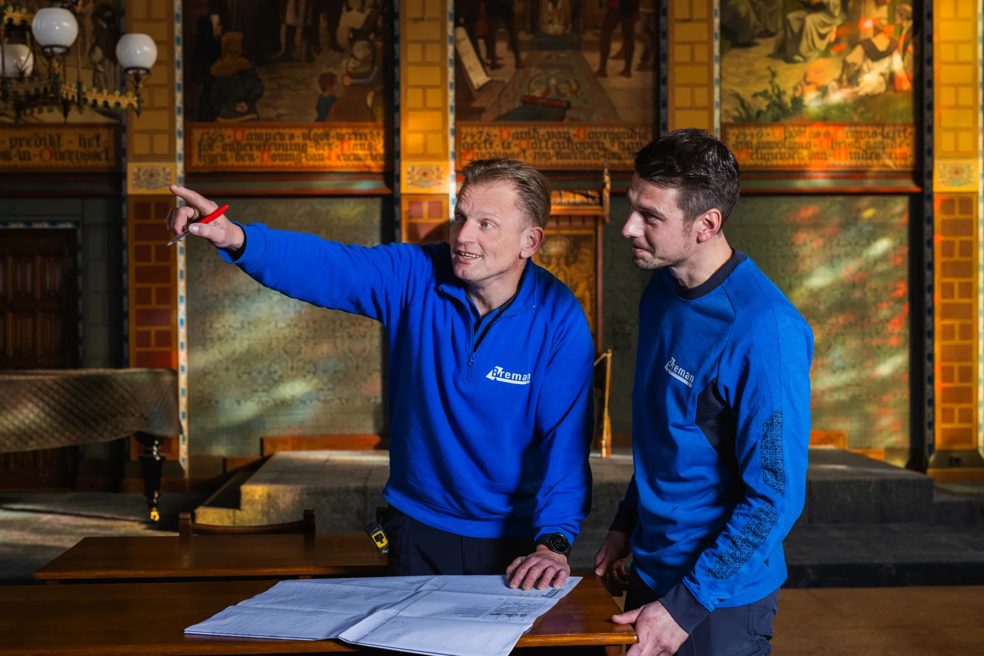 Twee Breman-collega's in blauwe werkkleding bekijken bouwtekeningen in een historische ruimte.