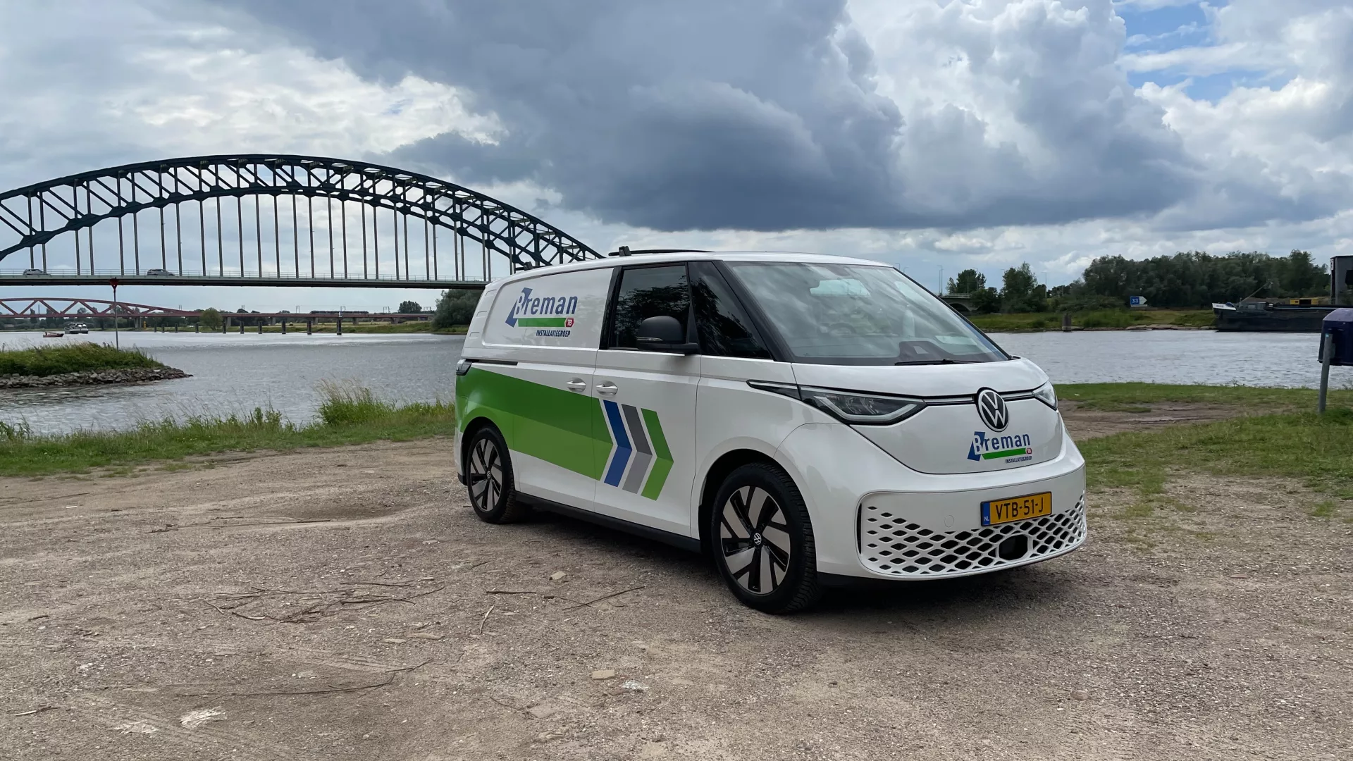 Elektrische Breman bus geparkeerd bij rivier met brug op de achtergrond.