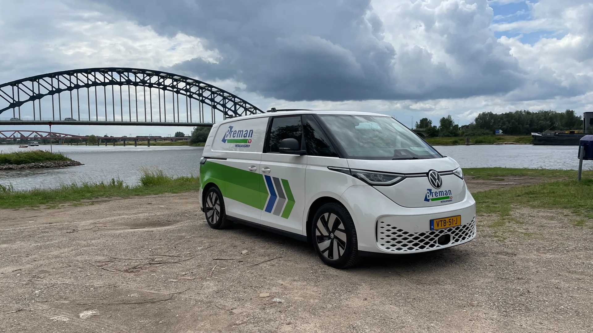 Elektrische Breman bus geparkeerd bij rivier met brug op de achtergrond.